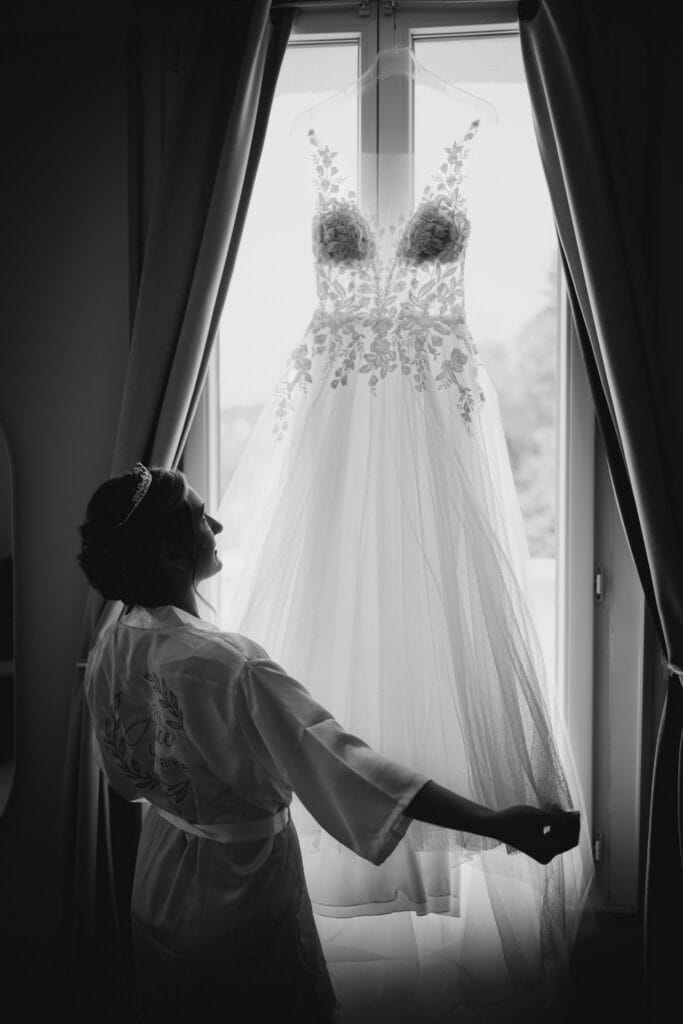 Mariée contemple sa robe suspendue à la fenêtre - Photographe mariage Versailles